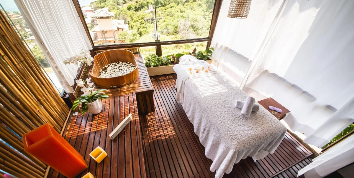 Cabine de spa com maca, banheira de madeira com flores e vista para a vegetação